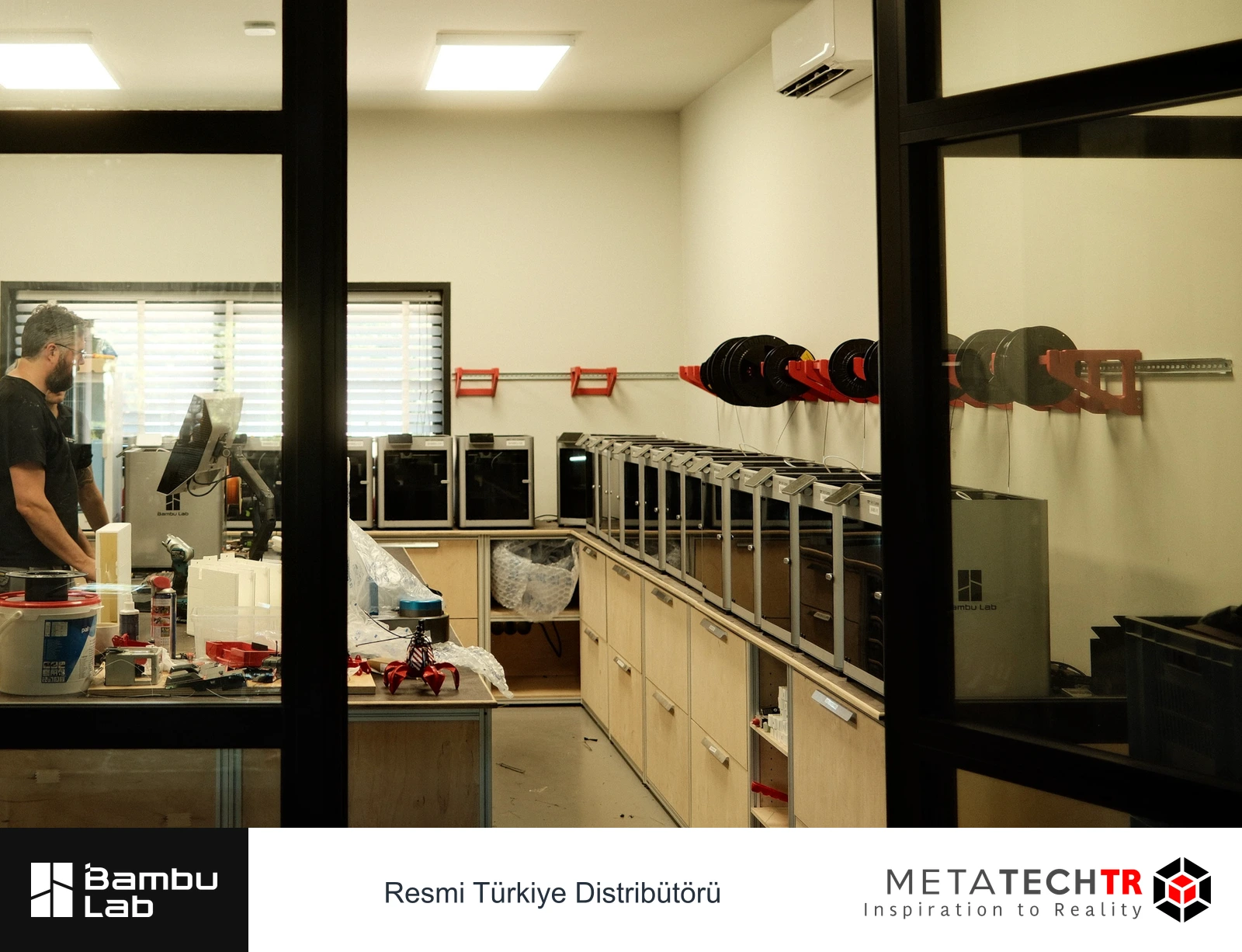 Cam bölme arkasında çalışma masası ve sıra hâlindeki yazıcılar.